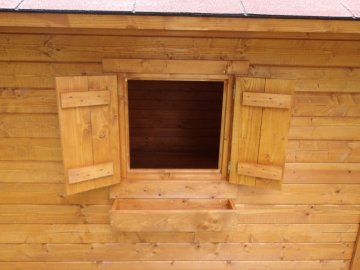 Dětský domeček Marii má okenní otvory vyplněné plexisklem, po stranách je doplněný dřevěnými okenicemi a pod oknem je připevněný truhlík na kytky